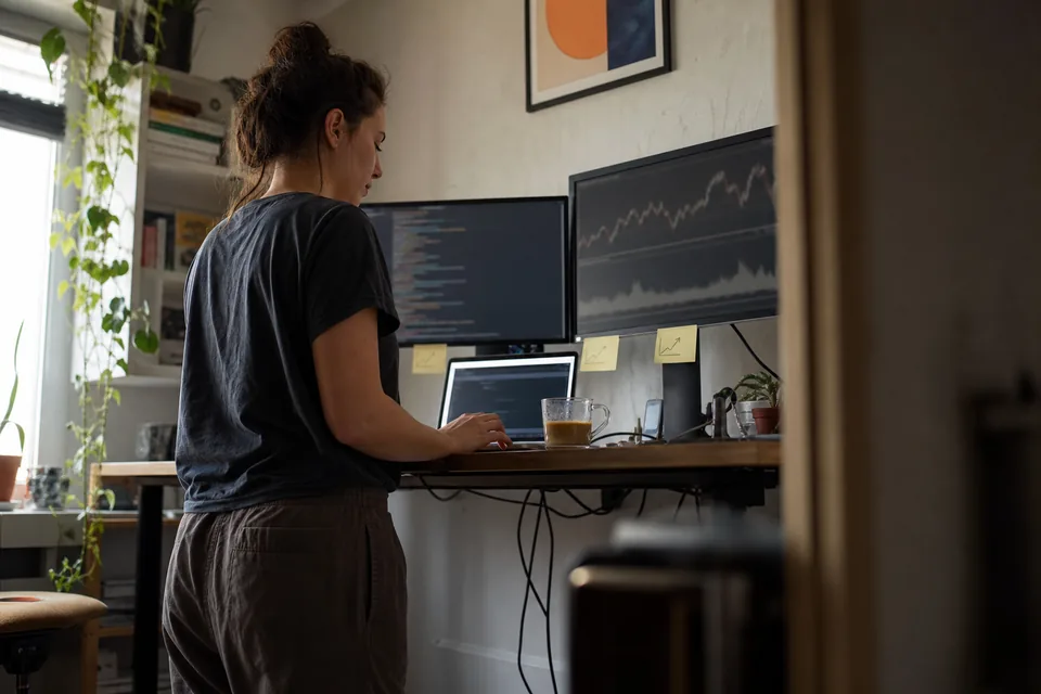 A developer at a standing desk with code on one monitor and a backtest equity curve on the other, sticky notes on the bezel