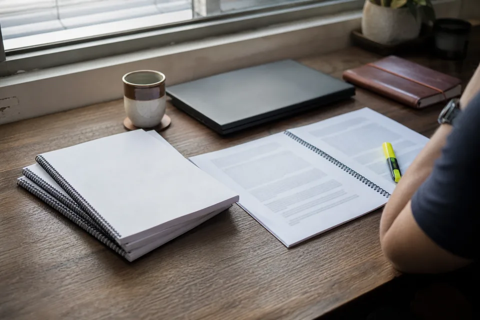 An investor reading a printed annual report with a highlighter at a desk by a window, beside a closed laptop and a notebook